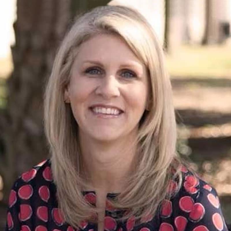 A woman with shoulder-length blonde hair is smiling outdoors. She is wearing a dark top with a red circular pattern, enjoying the sunshine after her shift at the emergency vet. Trees and sunlight are visible in the blurred background.