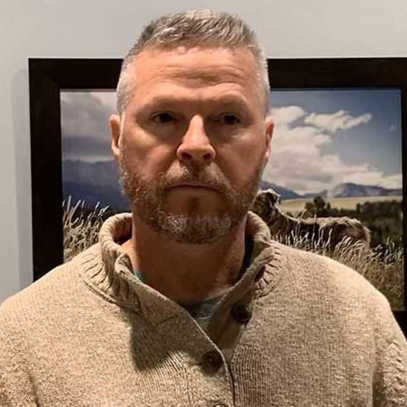 A man with short gray hair and a beard wearing a beige sweater stands indoors in front of a landscape photograph, perhaps at a Vet clinic or Pet ER, featuring grass, mountains, and a cloudy sky.