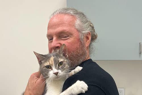 A man with gray hair and a beard hugs a gray and white cat in what appears to be an Emergency Vet clinic. The cat looks directly at the camera.