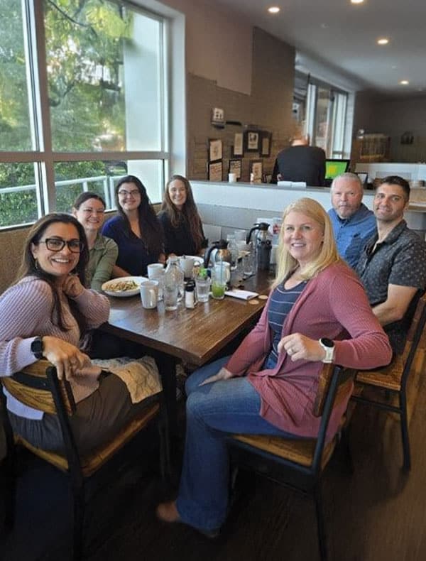 Seven people, including a veterinarian, sit around a wooden table in a bright, modern café, smiling at the camera. Food and drinks are on the table, and large windows let in natural light.