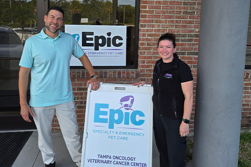 Two people stand smiling outside a building next to a sign that reads "Epic Specialty & Emergency Pet Care" and "Tampa Oncology Veterinary Cancer Center." A brick wall and glass door are in the background.