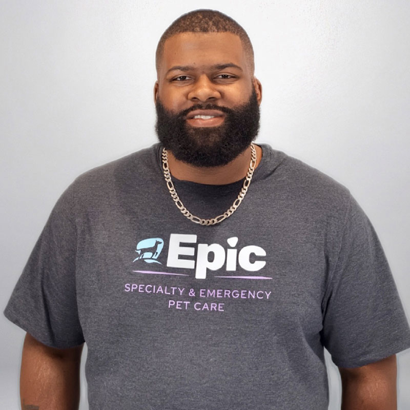 A bearded man wearing a gray "Epic Specialty & Emergency Pet Care" t-shirt and a silver chain necklace stands smiling in front of a light gray wall.