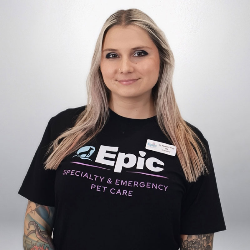 A smiling woman with long blonde hair and tattoos wears a black "Epic Specialty & Emergency Pet Care" T-shirt and a name tag, standing against a plain light background.