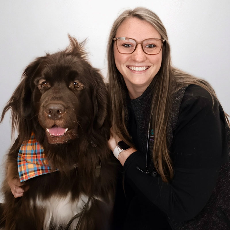 A smiling woman with long, straight hair and glasses poses next to a large, fluffy brown dog wearing a colorful plaid bandana against a plain light background.