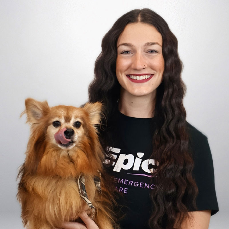A woman with long wavy brown hair smiles while holding a small, fluffy, brown dog that is licking its nose. The woman is wearing a black shirt with white text. Both are facing the camera against a plain background.