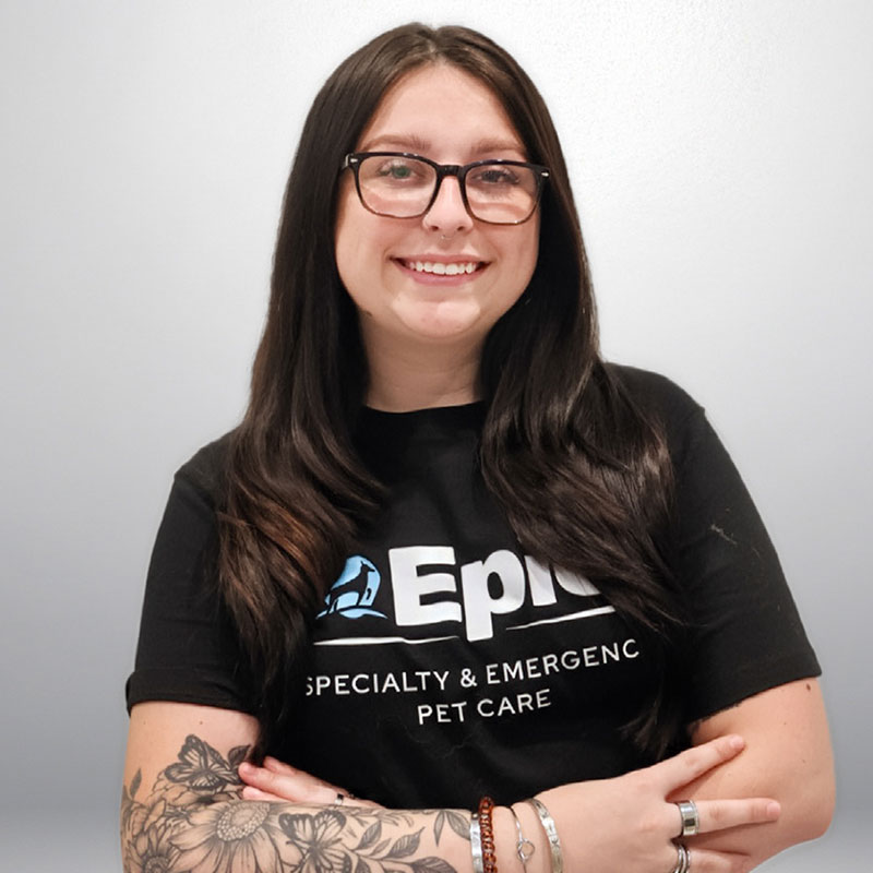 A woman with long dark hair and glasses smiles at the camera. She wears a black "Epic Specialty & Emergency Pet Care" t-shirt and has tattoos on her forearm. Her arms are crossed in front of her.