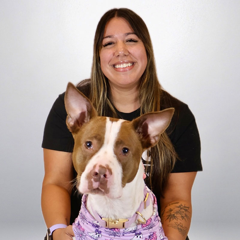 A smiling woman with long brown hair and a tattoo on her forearm poses with a brown and white dog wearing a pink patterned bandana. The background is plain and light-colored.