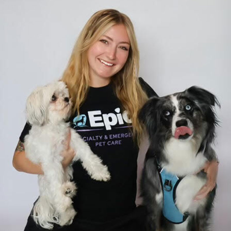 A smiling woman with long blonde hair holds a small white dog in one arm and stands next to a black and white dog wearing a blue harness. She is wearing a black "Epic Specialty & Emergency Pet Care" t-shirt.