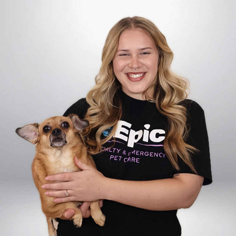 A smiling woman with long blonde hair wearing a black "Epic Pet Care" T-shirt holds a small brown dog with large ears in front of a plain light background.