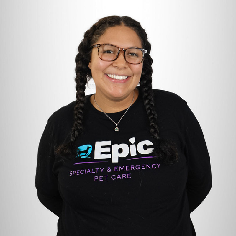 A person with glasses and braided hair smiles at the camera, wearing a black shirt that says "Epic Specialty & Emergency Pet Care" on a plain light background.