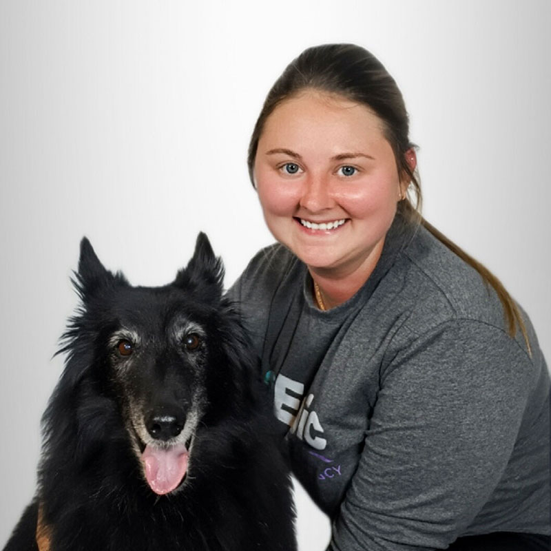 A smiling woman with brown hair in a ponytail, wearing a gray shirt, poses next to a large black dog with pointed ears and its tongue out, both in front of a plain light background.