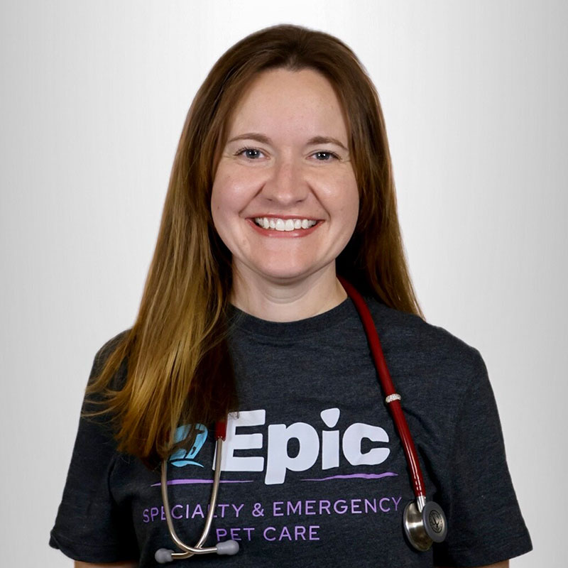A smiling woman with long brown hair wears a dark "Epic Specialty & Emergency Pet Care" T-shirt and has a red stethoscope draped around her neck, standing against a plain light background.
