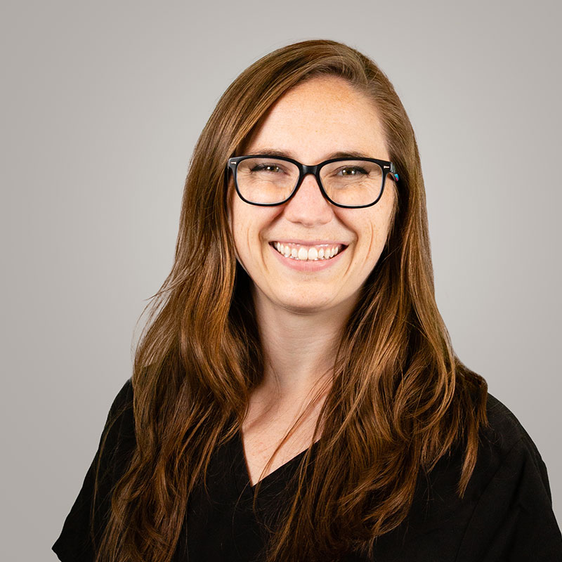 A woman with long brown hair and glasses smiles at the camera. She is wearing a black top and is posed against a plain, light gray background.