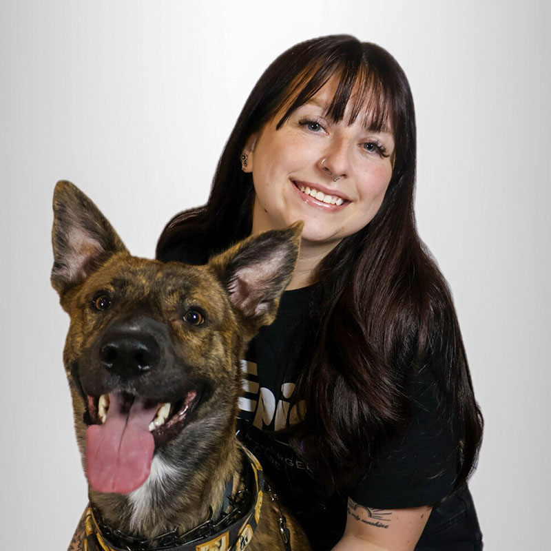 A smiling woman with long dark hair poses beside a happy, panting brown and black dog with upright ears against a plain white background.